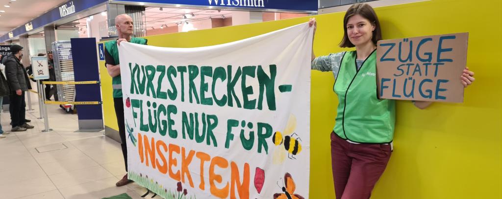 Aktive mit Protestbanner "Kurzstreckenflüge nur für Insekten" in der Abflughalle eines Flughafens