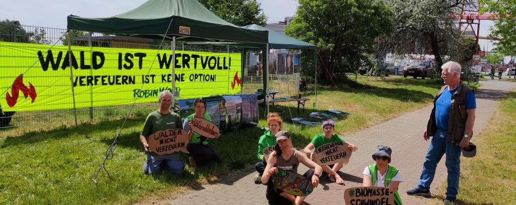 Aktive sitzen mit Protestschildern auf dem Boden vor einem Pelletwerk